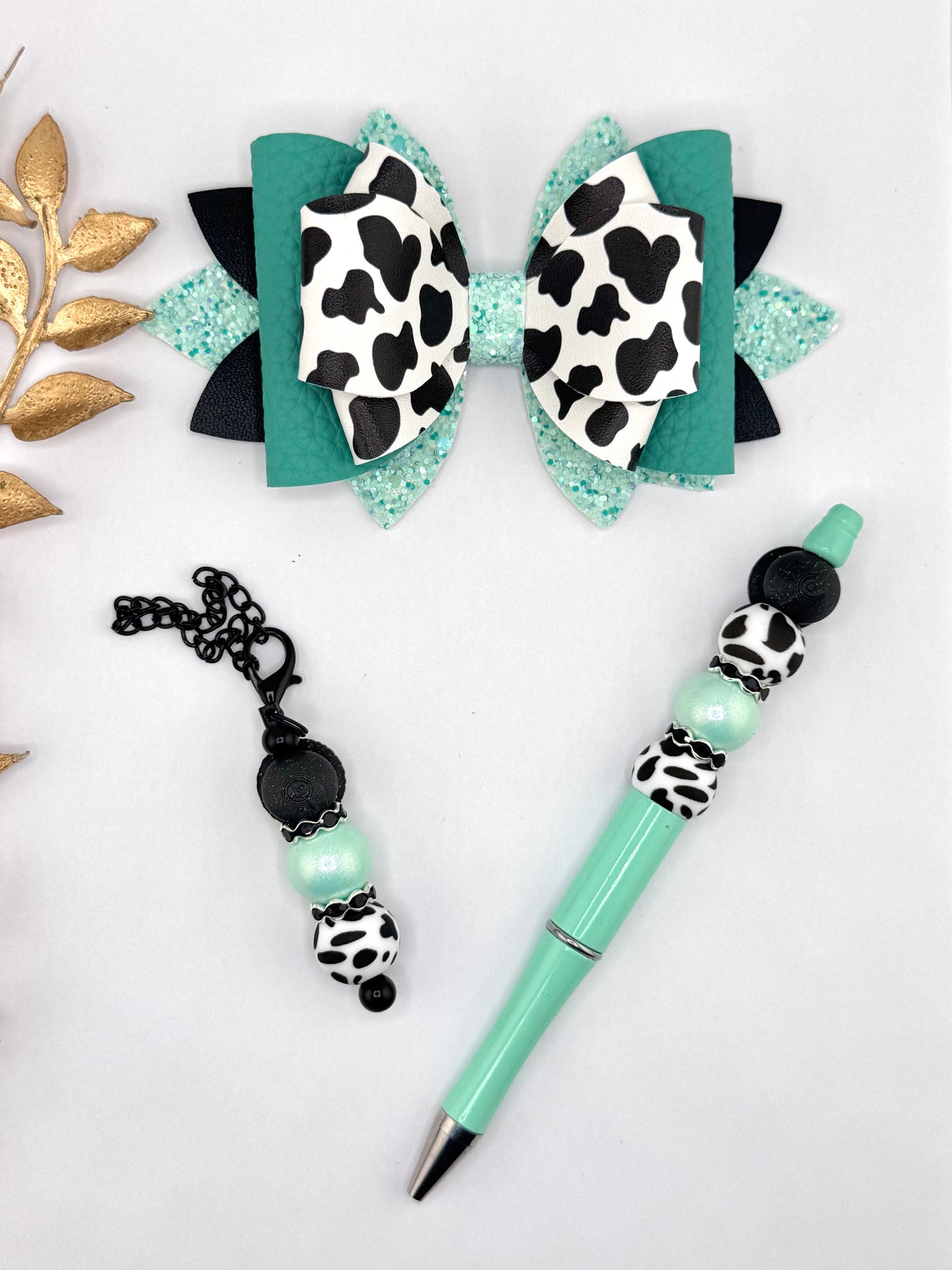 Teal faux leather bow set with cow print center and matching beaded pen and keychain charm in teal, aqua glitter, black, and white cow print beads.