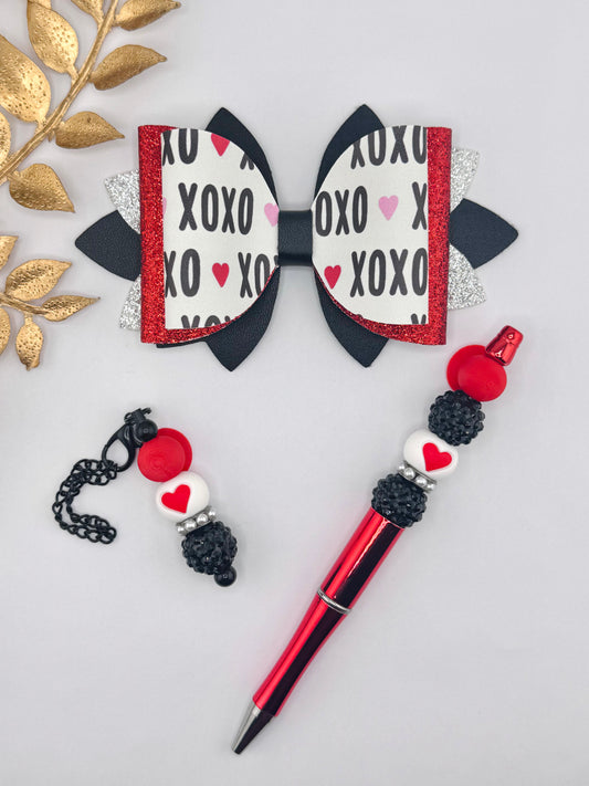 Valentine’s Day faux leather bow kit featuring an XOXO heart print bow layered with red glitter and black leather, paired with a red beaded pen and matching heart bead pen charm in red, black, and white.